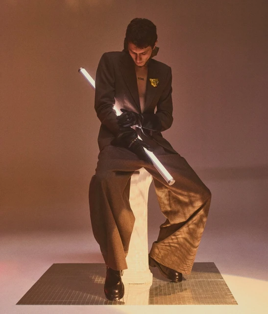 An adult male sits in a studio wearing a suit with wide-leg pants, black leather boots, and gloves, holding a light stick.
