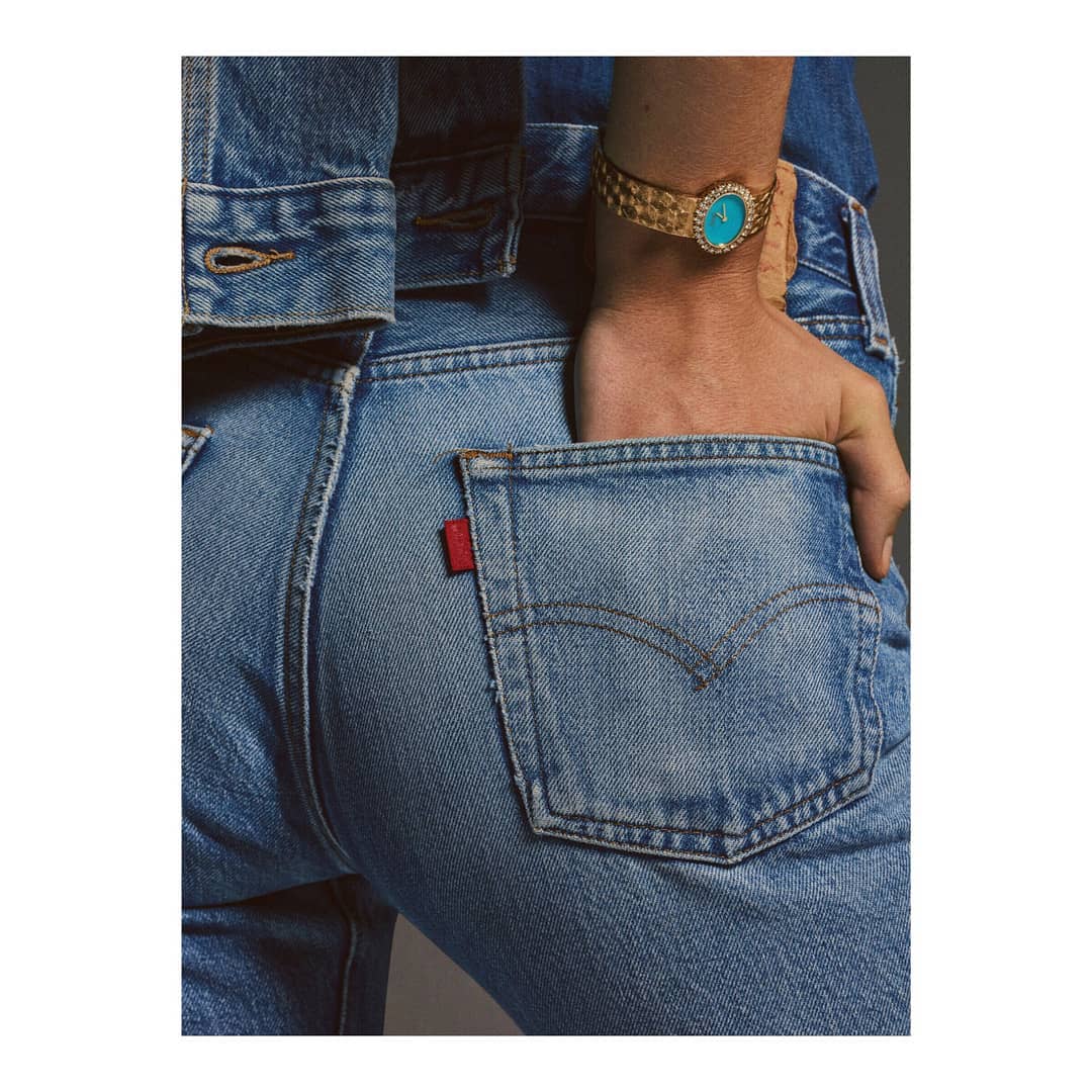Person in denim jacket and blue jeans with hand in back pocket displaying a gold watch with a turquoise face and bezel.