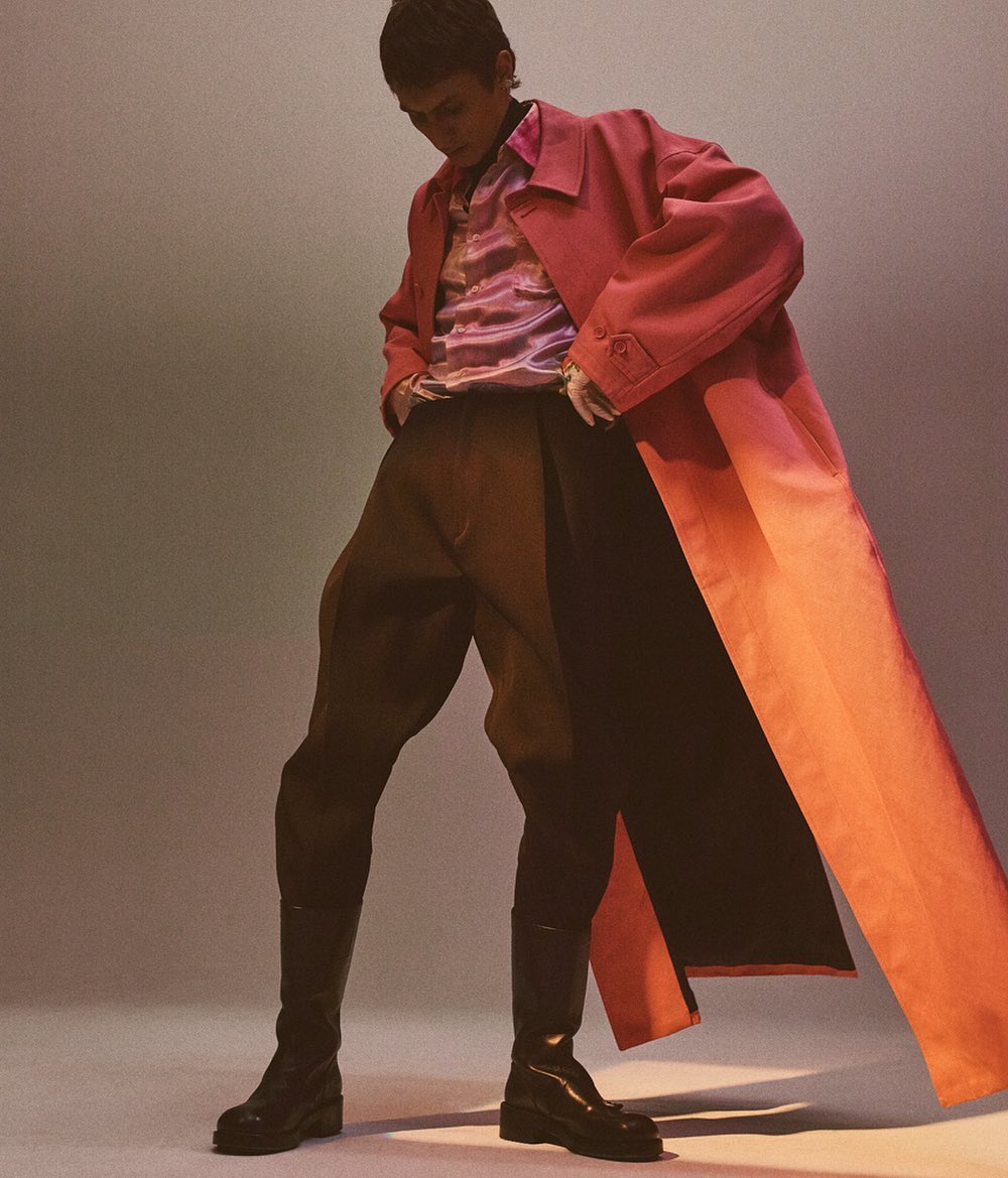 A fashion shot presents a slender adult posing in a salmon overcoat, dark pants, and shiny black boots against a gradient grey backdrop.