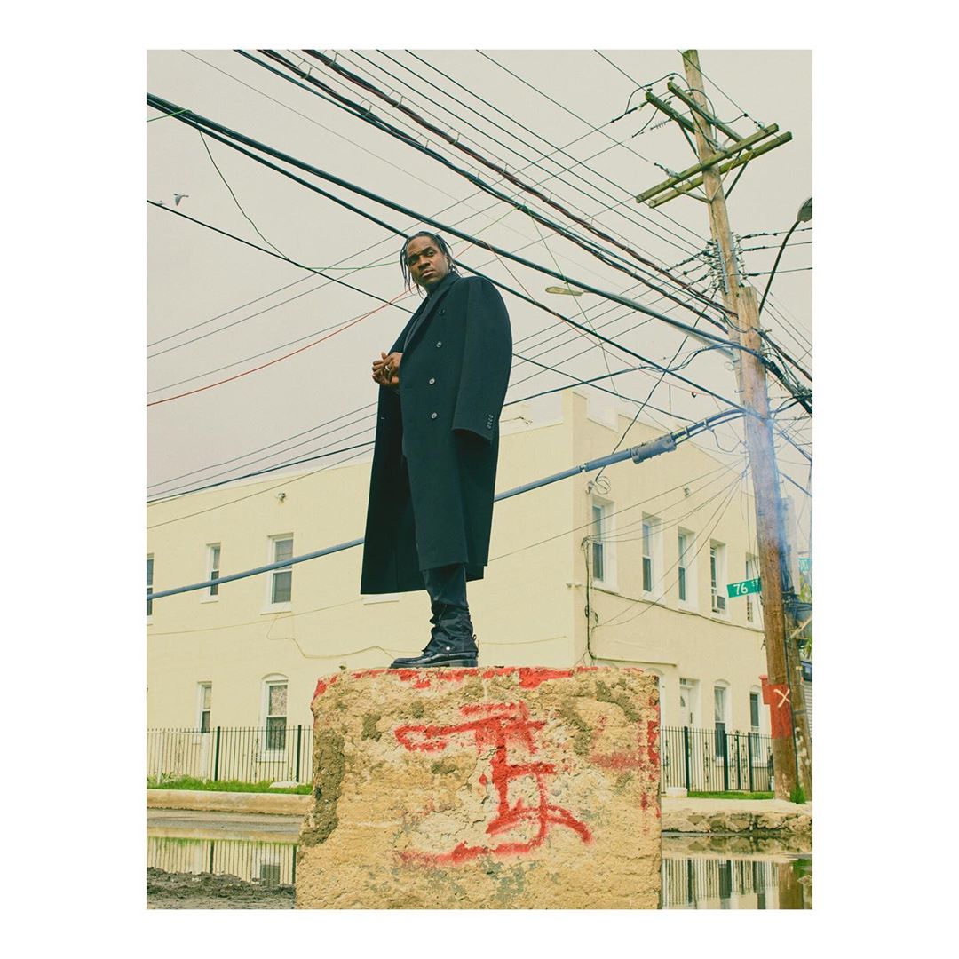 A man in a black coat stands atop a graffitied concrete block with utility poles and a building in the background on a cloudy day.