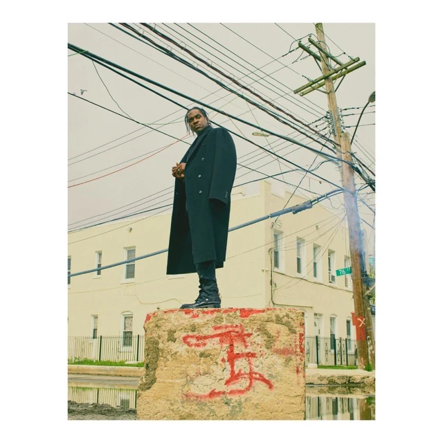 A man in a black coat stands atop a graffitied concrete block with utility poles and a building in the background on a cloudy day.