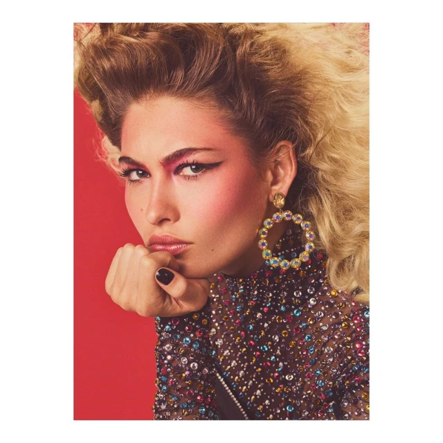 Close-up portrait of a stylish adult female with blonde hair and distinctive makeup, wearing jeweled earrings and a mesh top against red.