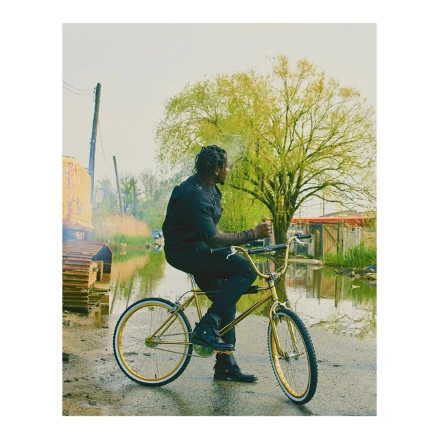 A man in black clothing rides a gold bicycle through a flooded street, near a construction vehicle, under a cloudy sky with green trees.