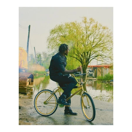 A man in black clothing rides a gold bicycle through a flooded street, near a construction vehicle, under a cloudy sky with green trees.