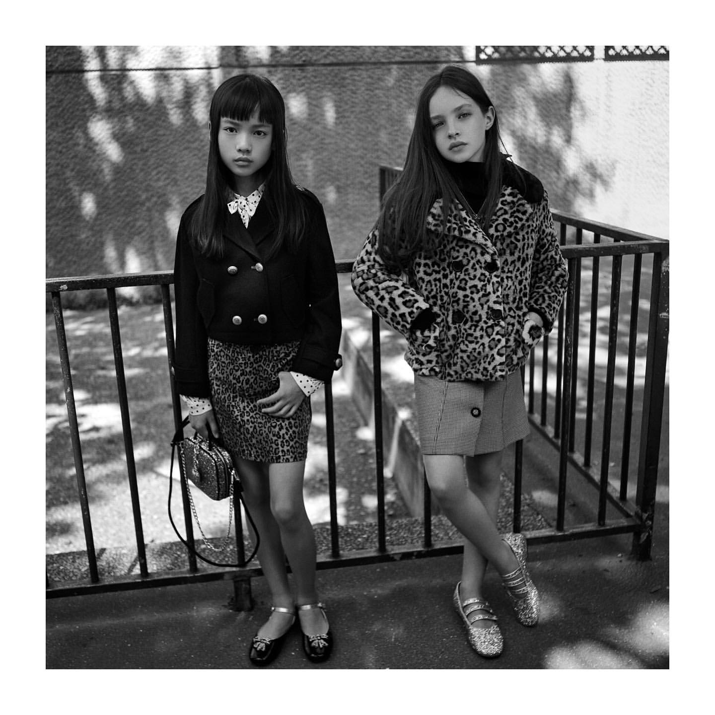 Two young girls in jackets and skirts stand in an urban outdoor setting, captured in black and white, showcasing fashion and style.