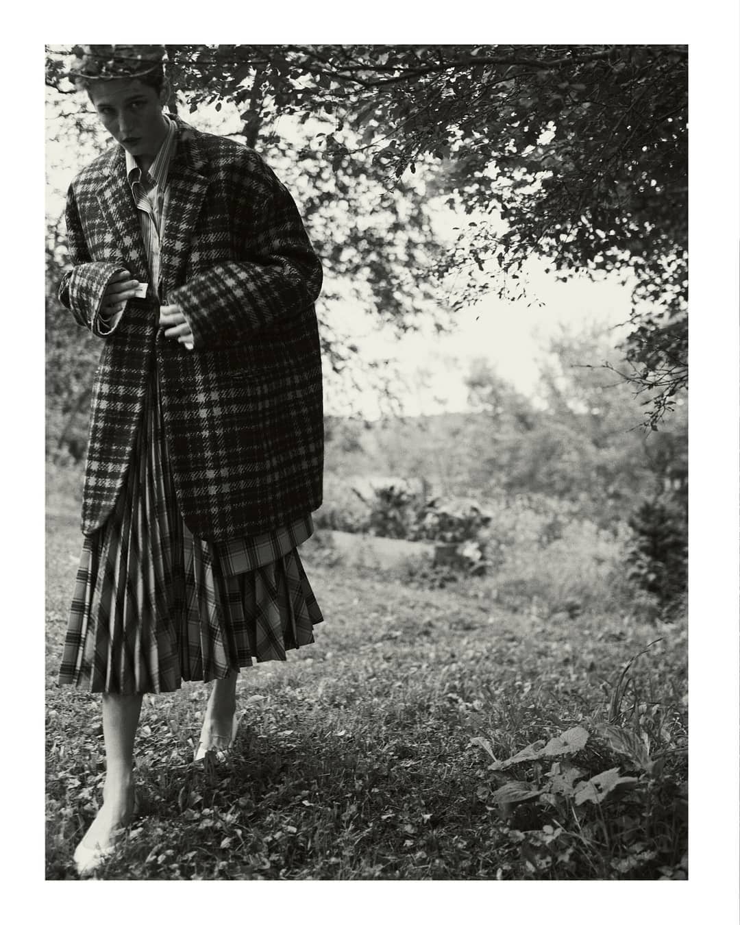 A person in a plaid coat and skirt stands in a grassy field under trees in a black and white fashion editorial shot.