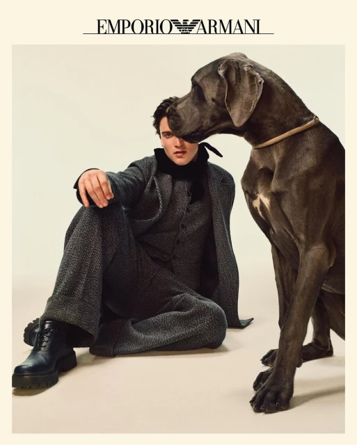 An adult male model in gray suit sitting indoors with a large gray Great Dane dog next to him in a studio fashion shot.