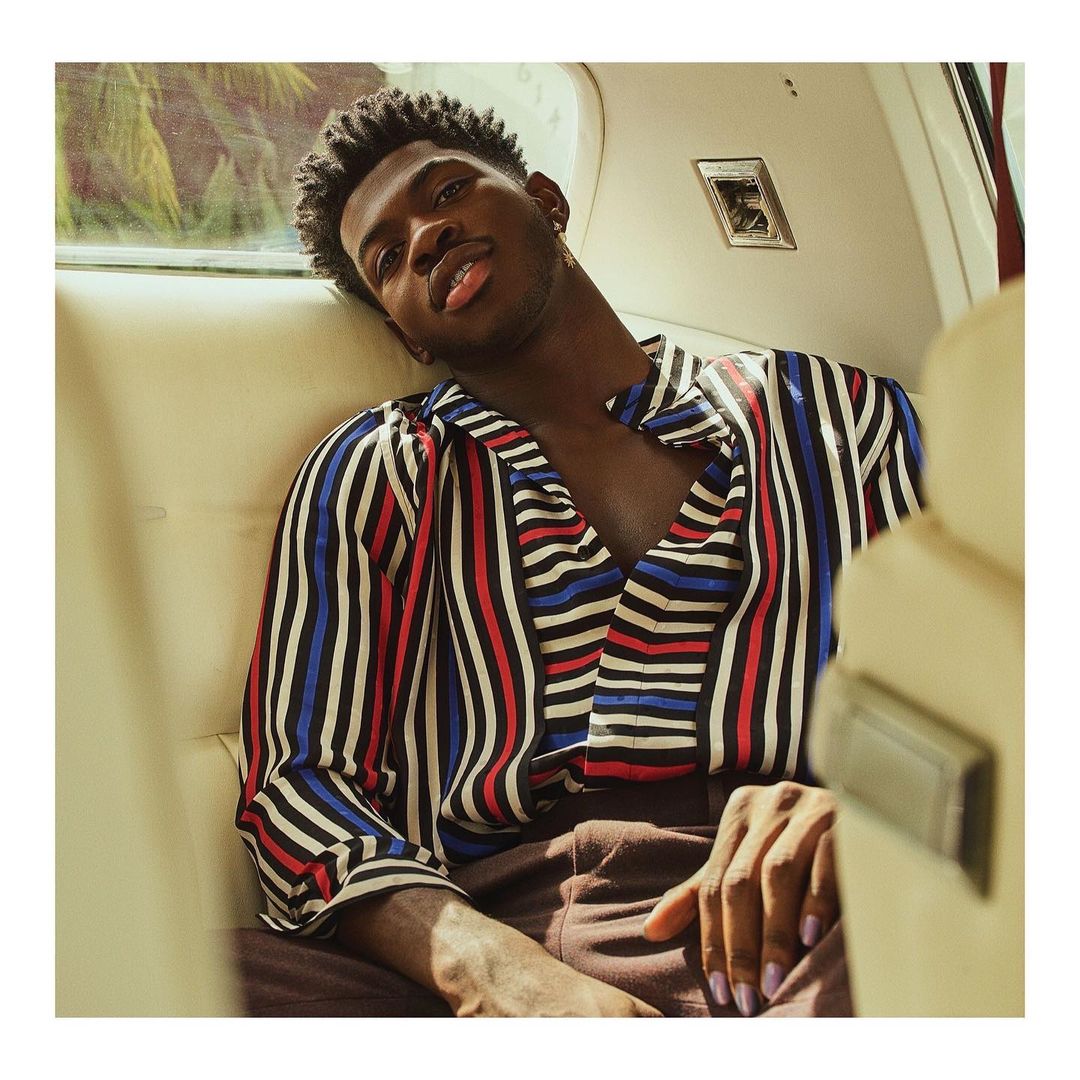 Close-up shot of a Black male in a vertical striped shirt, sitting inside an automobile, looking at the camera with a neutral expression.