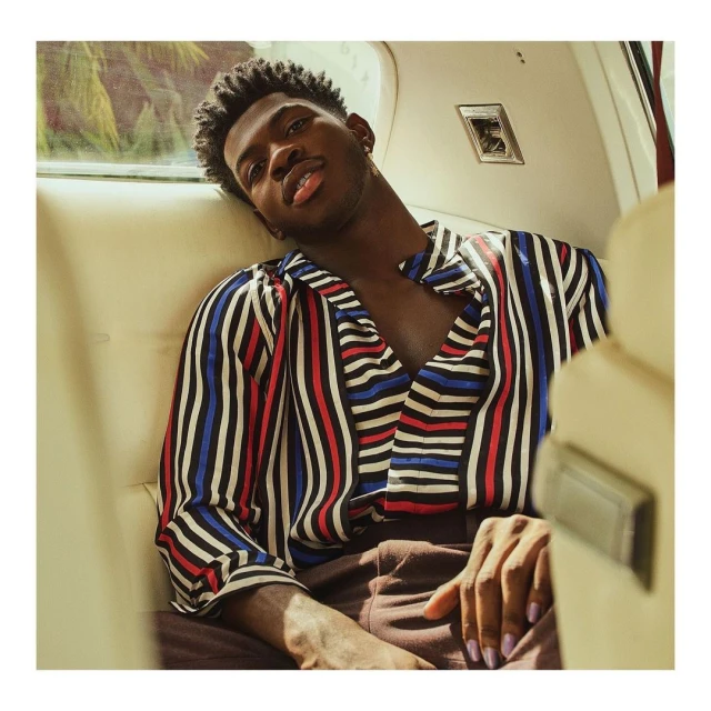 Close-up shot of a Black male in a vertical striped shirt, sitting inside an automobile, looking at the camera with a neutral expression.