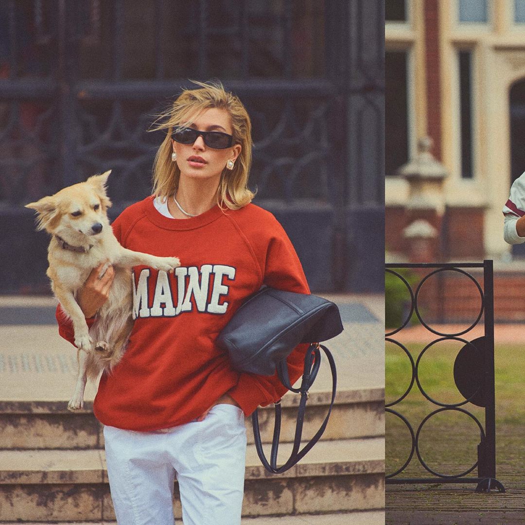 Blonde woman in red sweatshirt holds small dog in outdoor setting with building, wearing sunglasses and white pants.
