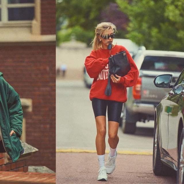 A blonde woman walks on a street wearing a red sweatshirt, black biker shorts, white sneakers and socks, sunglasses and carrying a bag.