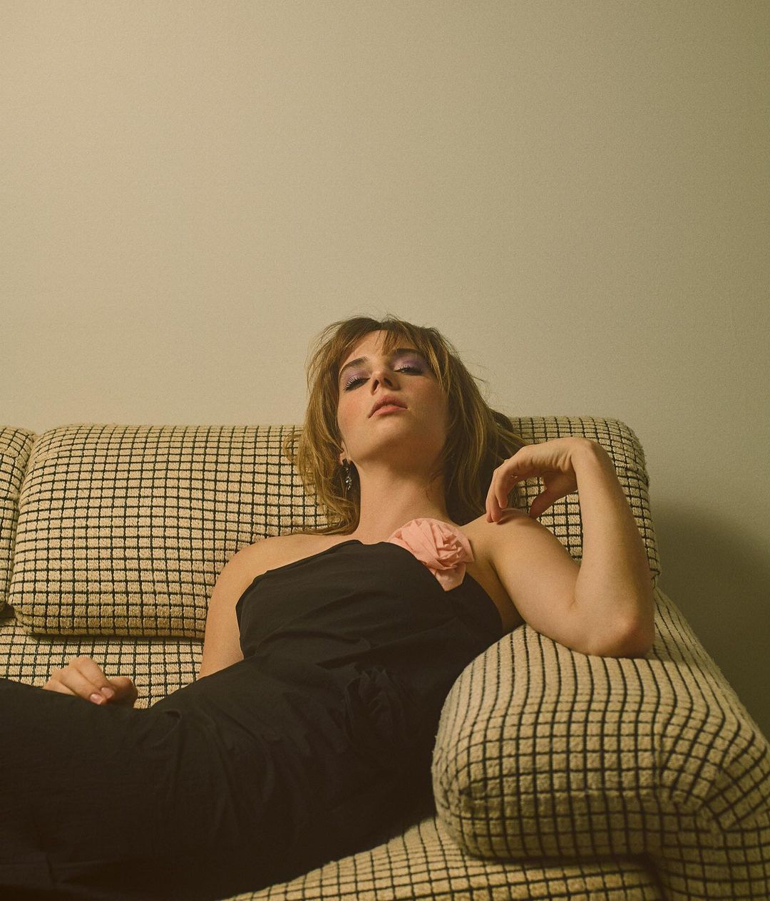Attractive woman in a black one-shoulder dress lounges on a checkered couch, looking at the camera with a neutral expression.