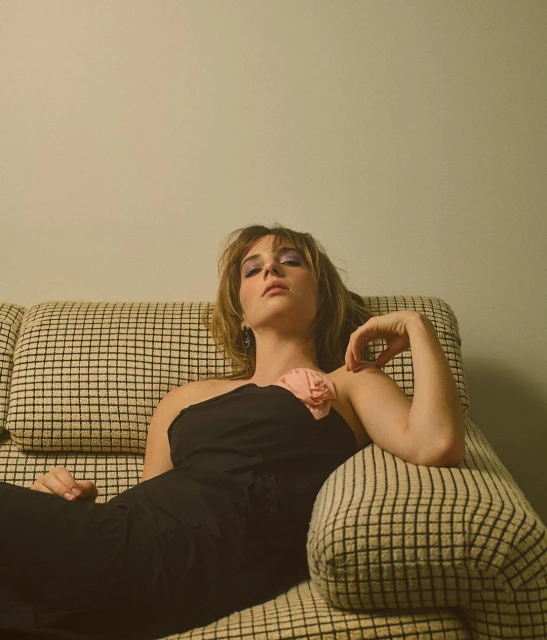 Attractive woman in a black one-shoulder dress lounges on a checkered couch, looking at the camera with a neutral expression.