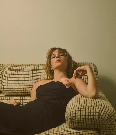 Attractive woman in a black one-shoulder dress lounges on a checkered couch, looking at the camera with a neutral expression.