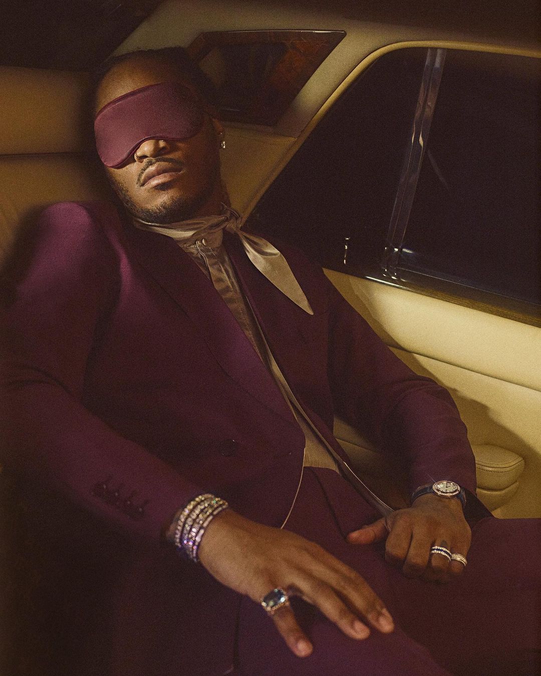 Man in a plum suit with a blindfold seated inside a car, wearing a watch, rings, and bracelets, creating a sophisticated, enigmatic portrait.