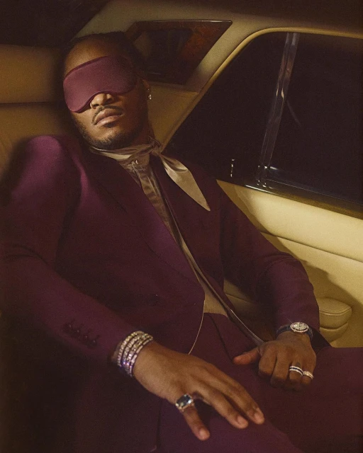 Man in a plum suit with a blindfold seated inside a car, wearing a watch, rings, and bracelets, creating a sophisticated, enigmatic portrait.