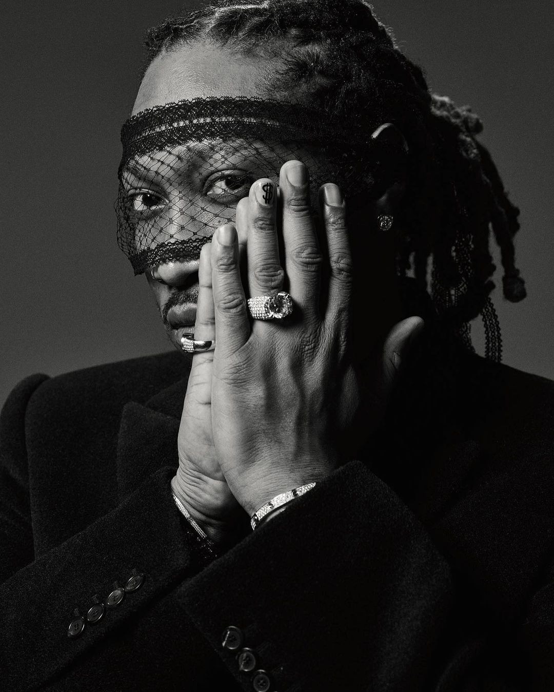 Monochrome portrait of a Black adult male with dreadlocks, wearing a veil and jacket, with rings and bracelets, hands near face. 