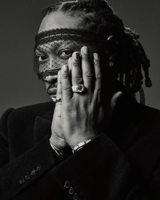 Monochrome portrait of a Black adult male with dreadlocks, wearing a veil and jacket, with rings and bracelets, hands near face. 