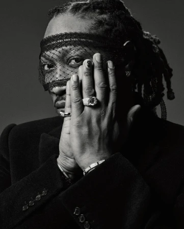 Monochrome portrait of a Black adult male with dreadlocks, wearing a veil and jacket, with rings and bracelets, hands near face. 