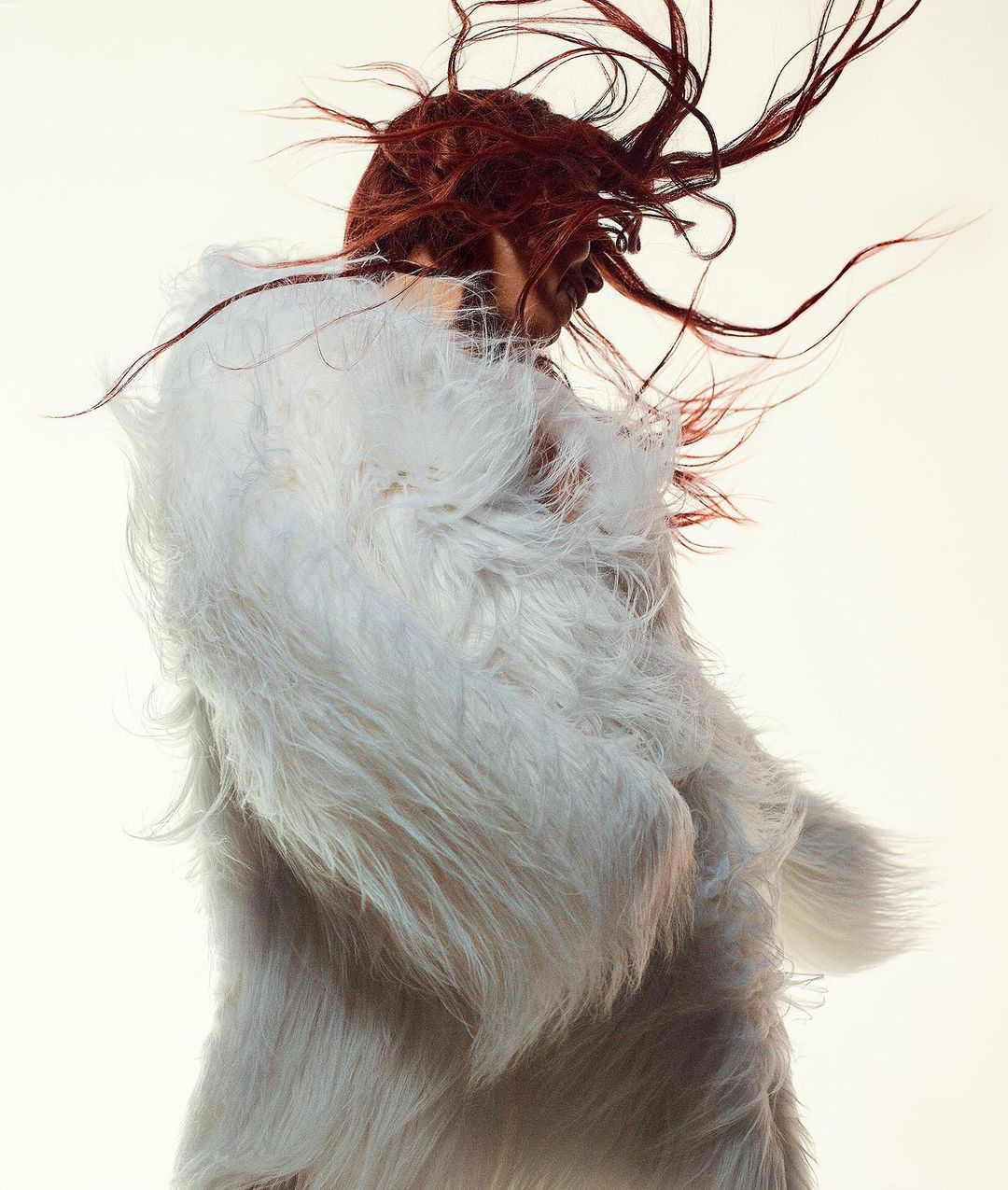 Woman in a white faux fur coat with flowing red hair against a bright white background. Fashion photography.