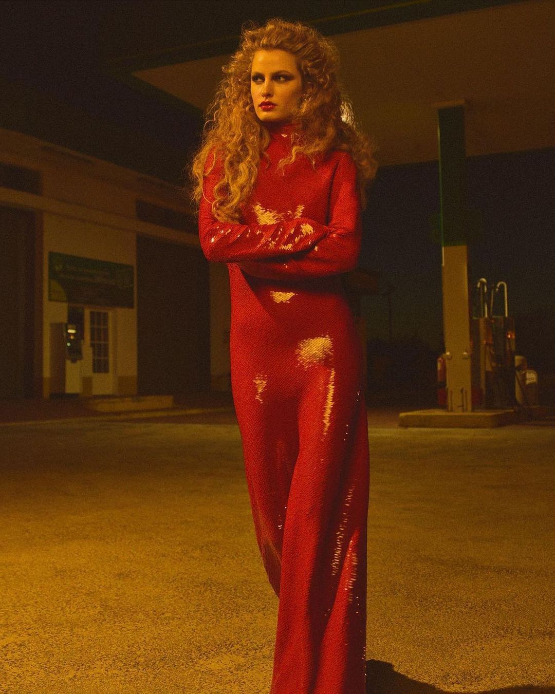 Adult woman poses in a red, sequined dress against the backdrop of a vintage gas station at night, illuminated by street lights.