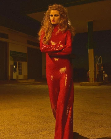 Adult woman poses in a red, sequined dress against the backdrop of a vintage gas station at night, illuminated by street lights.