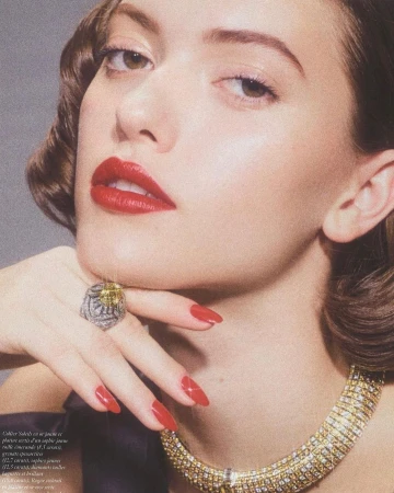 Close-up portrait of a woman with red lipstick and manicured nails, wearing a gold necklace and ring against a gray background.