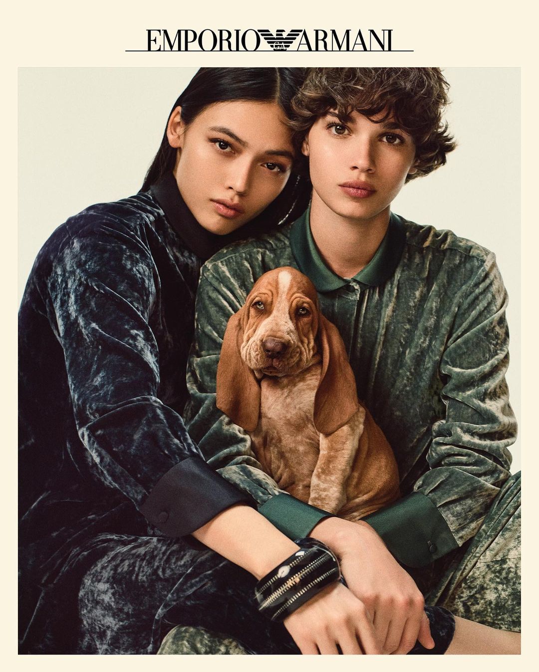 Fashion editorial featuring two models and a Basset Hound puppy, wearing green and blue velvet outfits by Emporio Armani against a light backdrop.