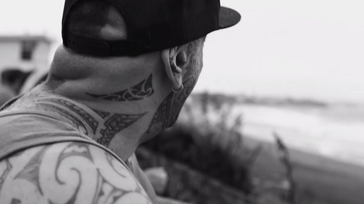 Monochrome portrait of a tattooed adult male wearing a cap, looking at a blurred sea and beachside landscape in the distance.