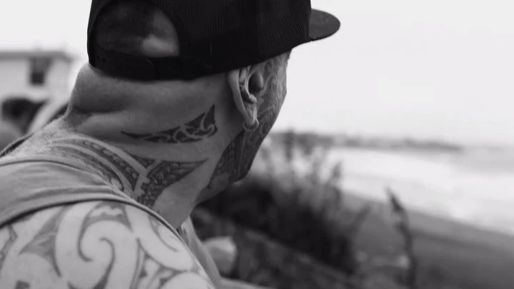 Monochrome portrait of a tattooed adult male wearing a cap, looking at a blurred sea and beachside landscape in the distance.