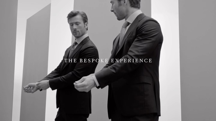 Black and white shot of a man in a dark suit adjusting his sleeve in front of a mirror, promoting a bespoke experience.