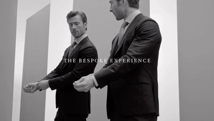 Black and white shot of a man in a dark suit adjusting his sleeve in front of a mirror, promoting a bespoke experience.