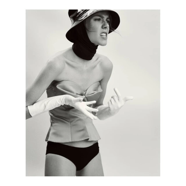 A woman in a hat and vintage bathing suit poses in a photo studio with white gloves and an angry facial expression.