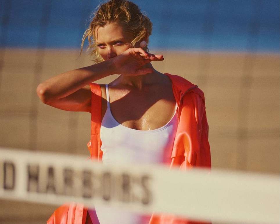 A woman in a white swimsuit and an orange jacket stands on a beach near a volleyball net with blue ocean and sky in the background.