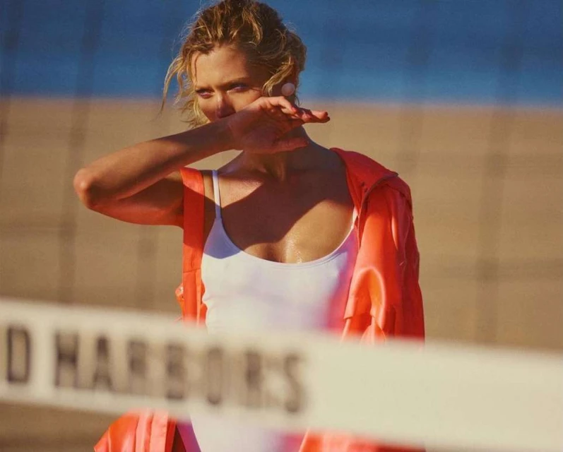 A woman in a white swimsuit and an orange jacket stands on a beach near a volleyball net with blue ocean and sky in the background.