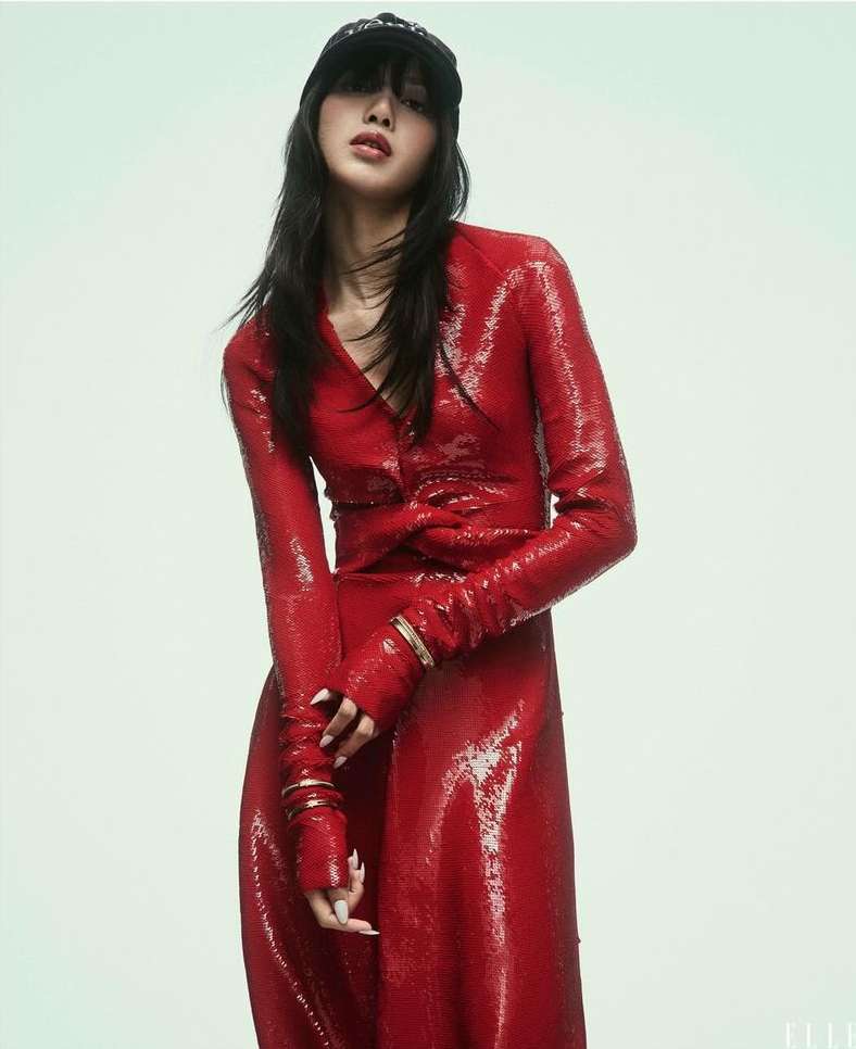 Woman in shiny red dress and black cap posing in photo studio, portrait orientation, fashion editorial, neutral expression. 