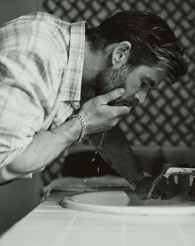 Monochrome portrait of an adult male washing his face over a bathroom sink, wearing a plaid shirt with water splashing.