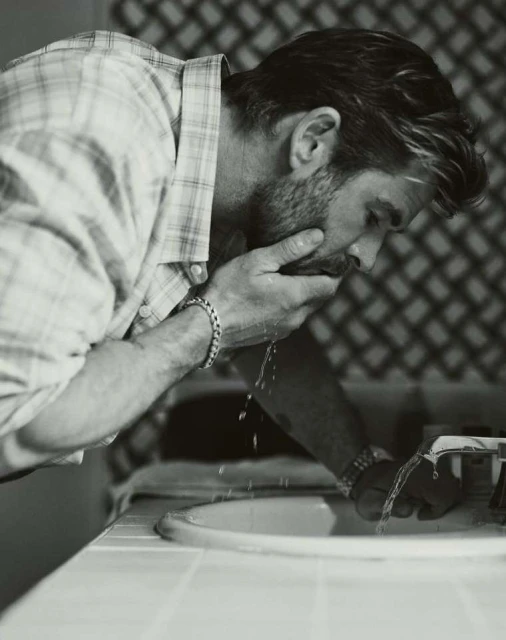 Monochrome portrait of an adult male washing his face over a bathroom sink, wearing a plaid shirt with water splashing.