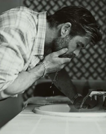 Monochrome portrait of an adult male washing his face over a bathroom sink, wearing a plaid shirt with water splashing.