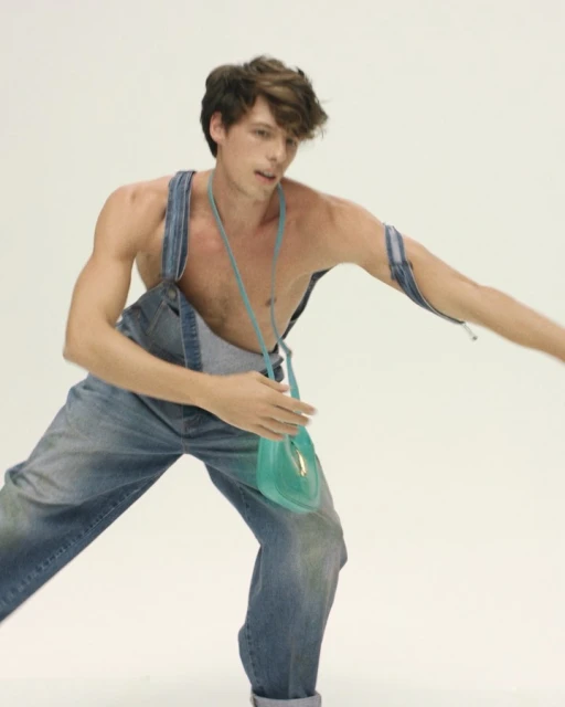 Man in denim overalls poses in a studio with a teal purse. He is shirtless and looking to the left with a slight smile on his face.