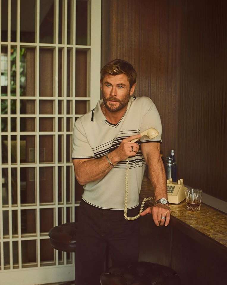 Chris Hemsworth, a white adult male, poses indoors holding a rotary telephone in a retro-styled setting with a glass of whiskey.