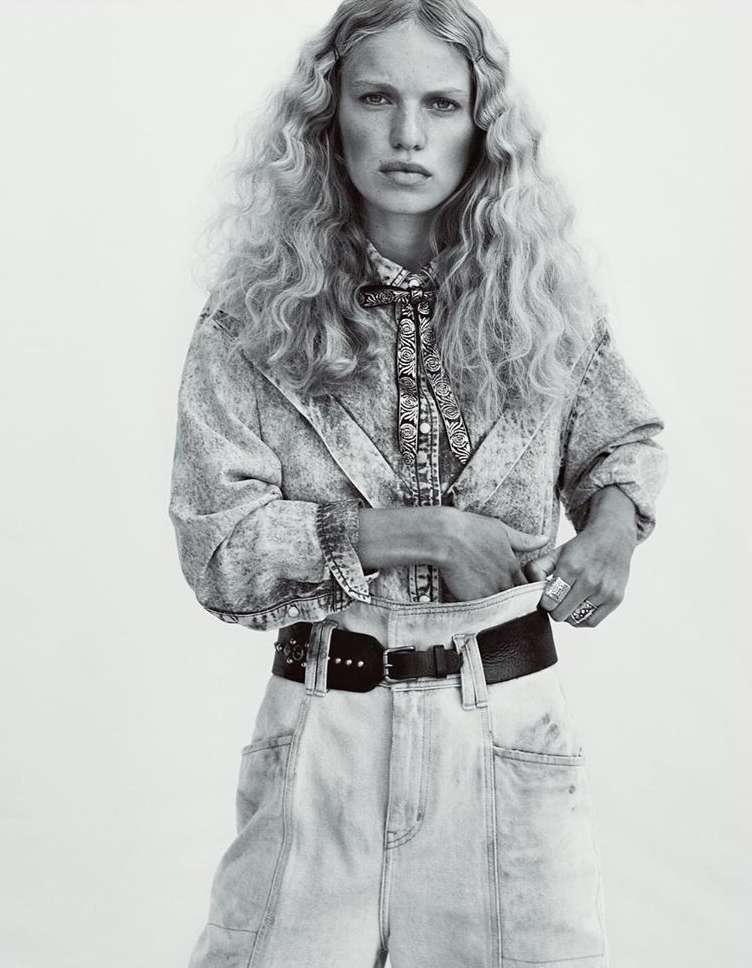A woman with wavy blonde hair poses in a black-and-white studio shot, wearing a denim outfit with a patterned shirt and a bolo tie.