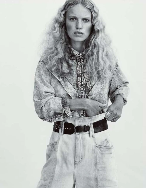 A woman with wavy blonde hair poses in a black-and-white studio shot, wearing a denim outfit with a patterned shirt and a bolo tie.