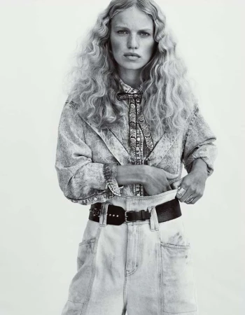 A woman with wavy blonde hair poses in a black-and-white studio shot, wearing a denim outfit with a patterned shirt and a bolo tie.