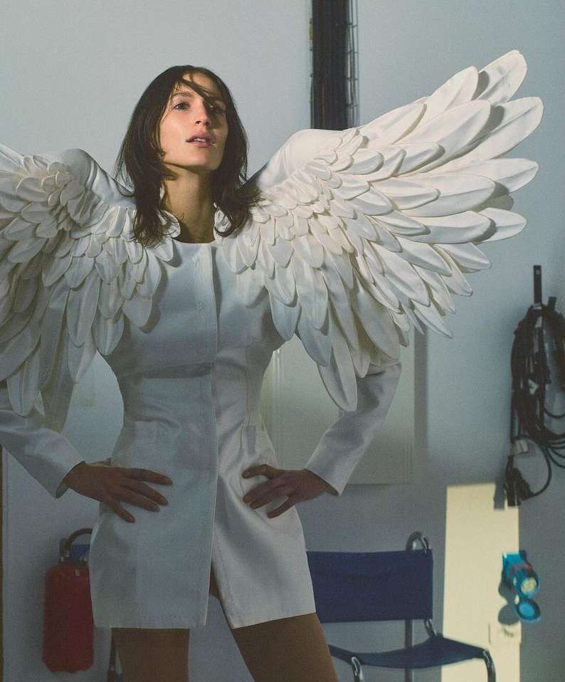 Woman in white mini dress with angel wings in studio, hands on hips, looking at camera. Modern take on angelic fashion.