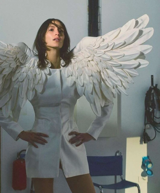 Woman in white mini dress with angel wings in studio, hands on hips, looking at camera. Modern take on angelic fashion.