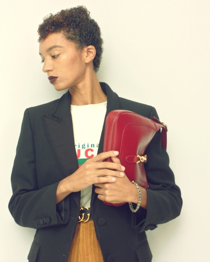 Model with short curly brown hair wearing a black blazer and holding a red handbag by photographer Gregory Harris
