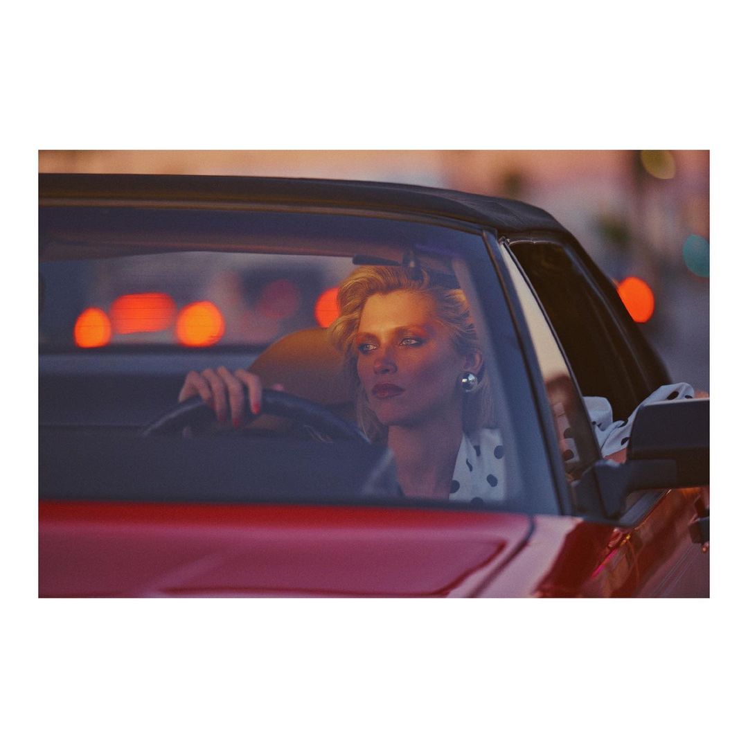 A stylish woman driving a red convertible car in an urban setting at dusk, capturing a fashionable and vintage aesthetic.