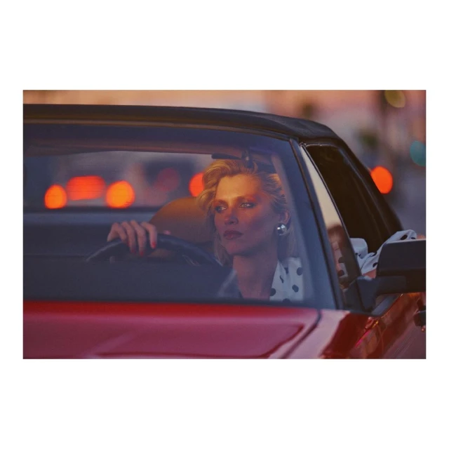 A stylish woman driving a red convertible car in an urban setting at dusk, capturing a fashionable and vintage aesthetic.