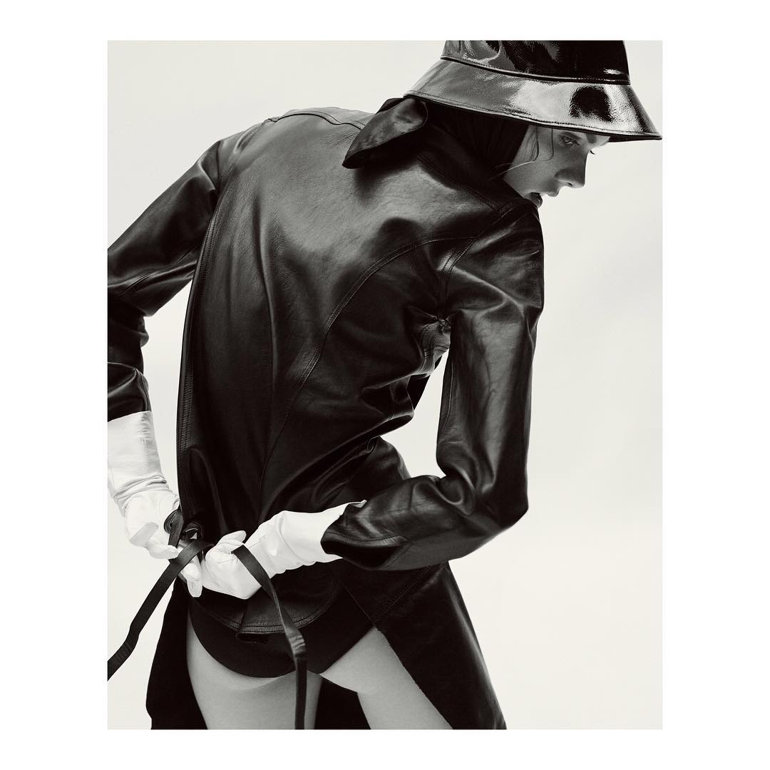 Monochrome studio fashion shot of a model in a black leather coat, bucket hat, and white gloves posing with a dark belt around her waist.